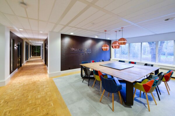 Vergaderruimte in Ypsilon Park Lounge met grote houten tafel, kleurrijke stoelen en koperen hanglampen.